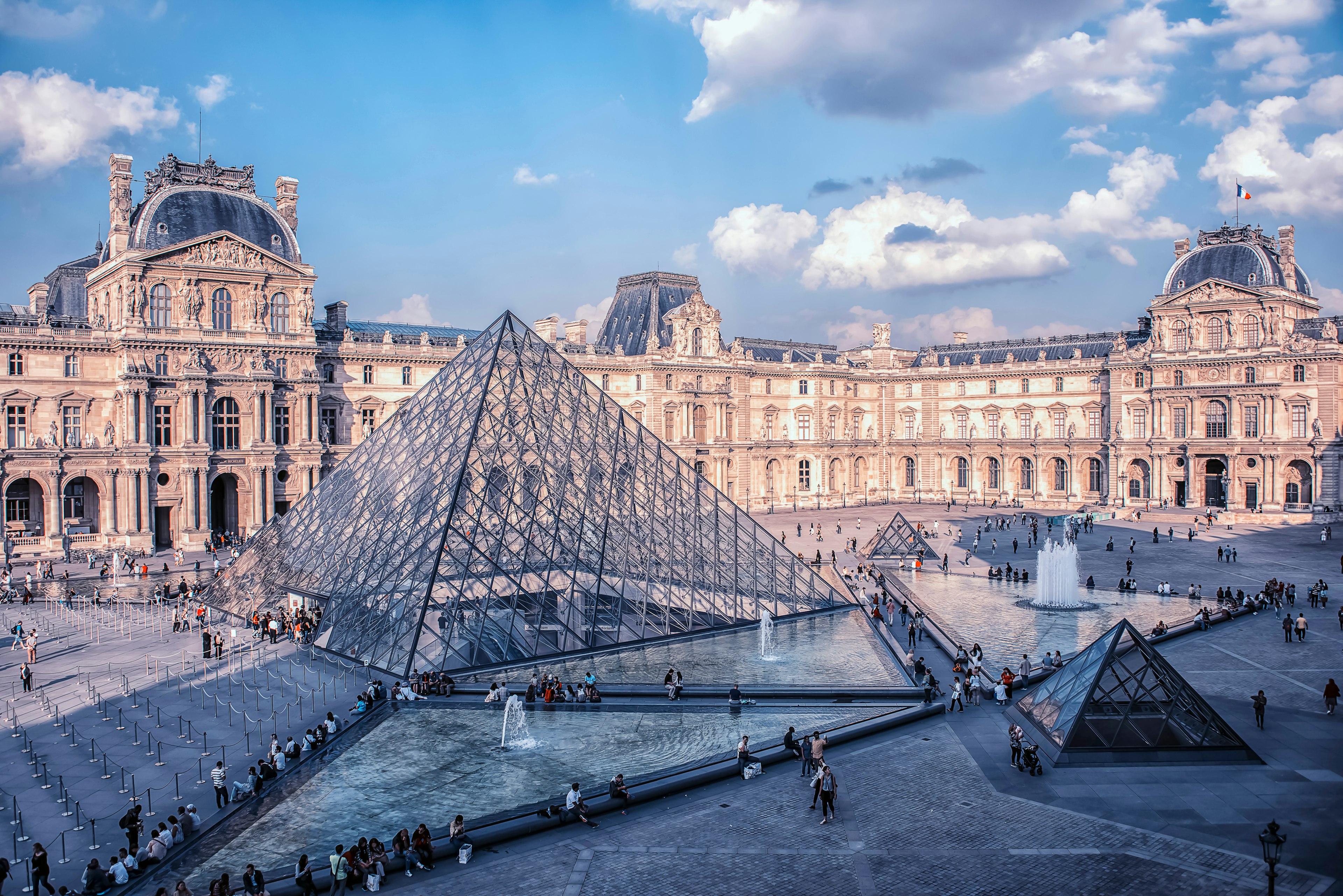 People walk around outside the Louvre museum in daytime.