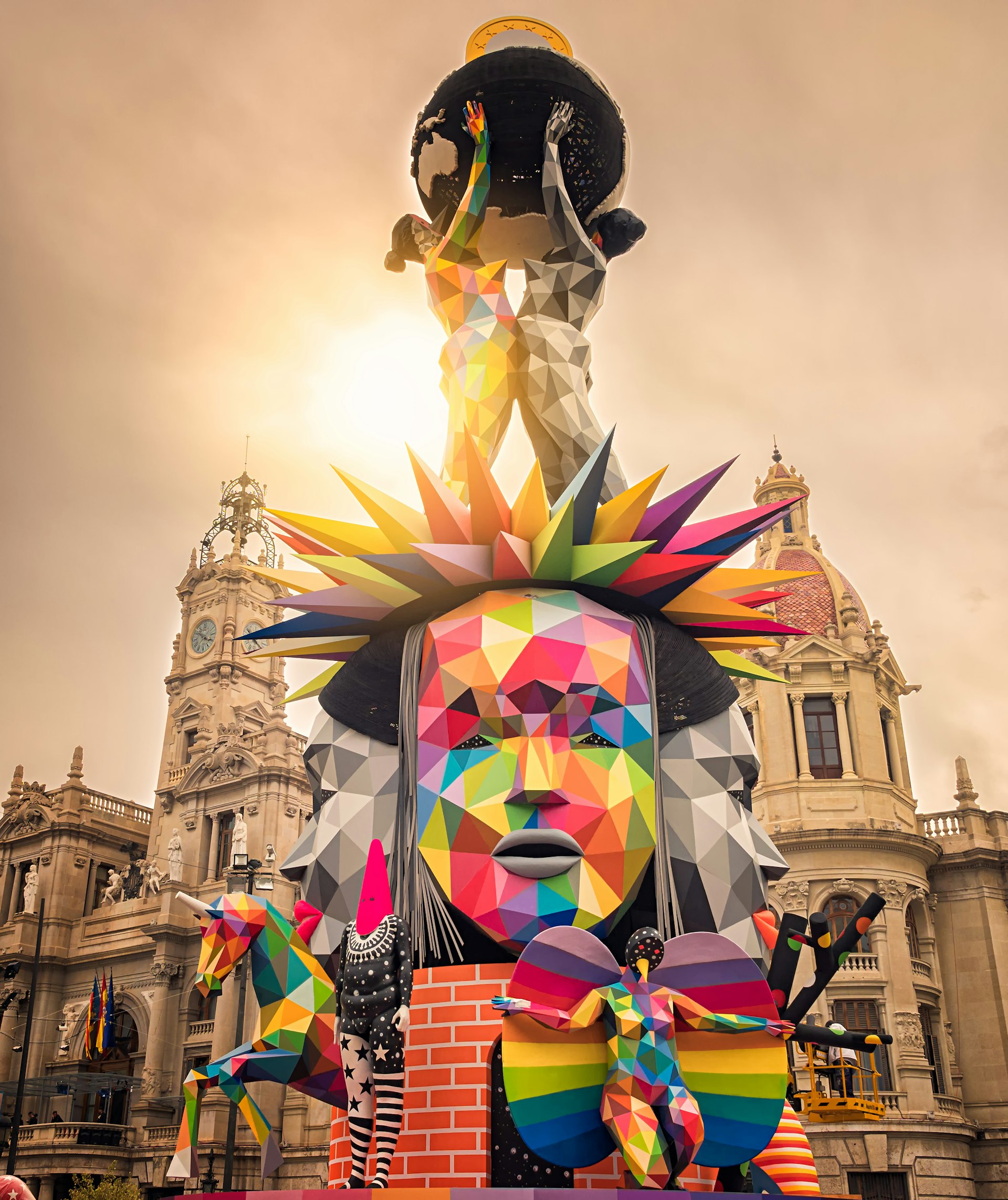 A huge figurative sculpture with several large faces at the center. It's made up of different colored geometric shapes.