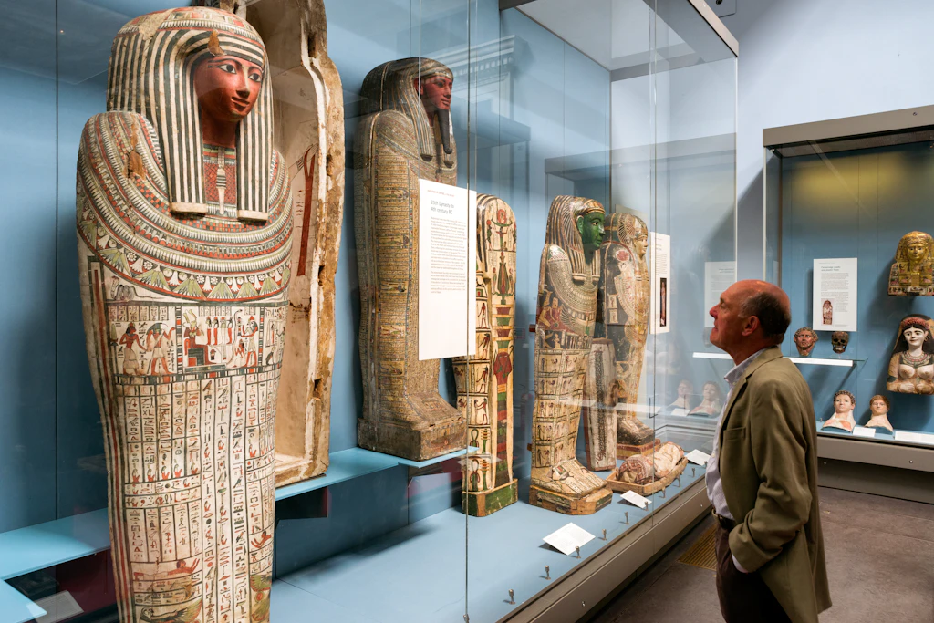 A set of Egyptian artefacts in a display case in a museum. An older white man in a suit is admiring them.