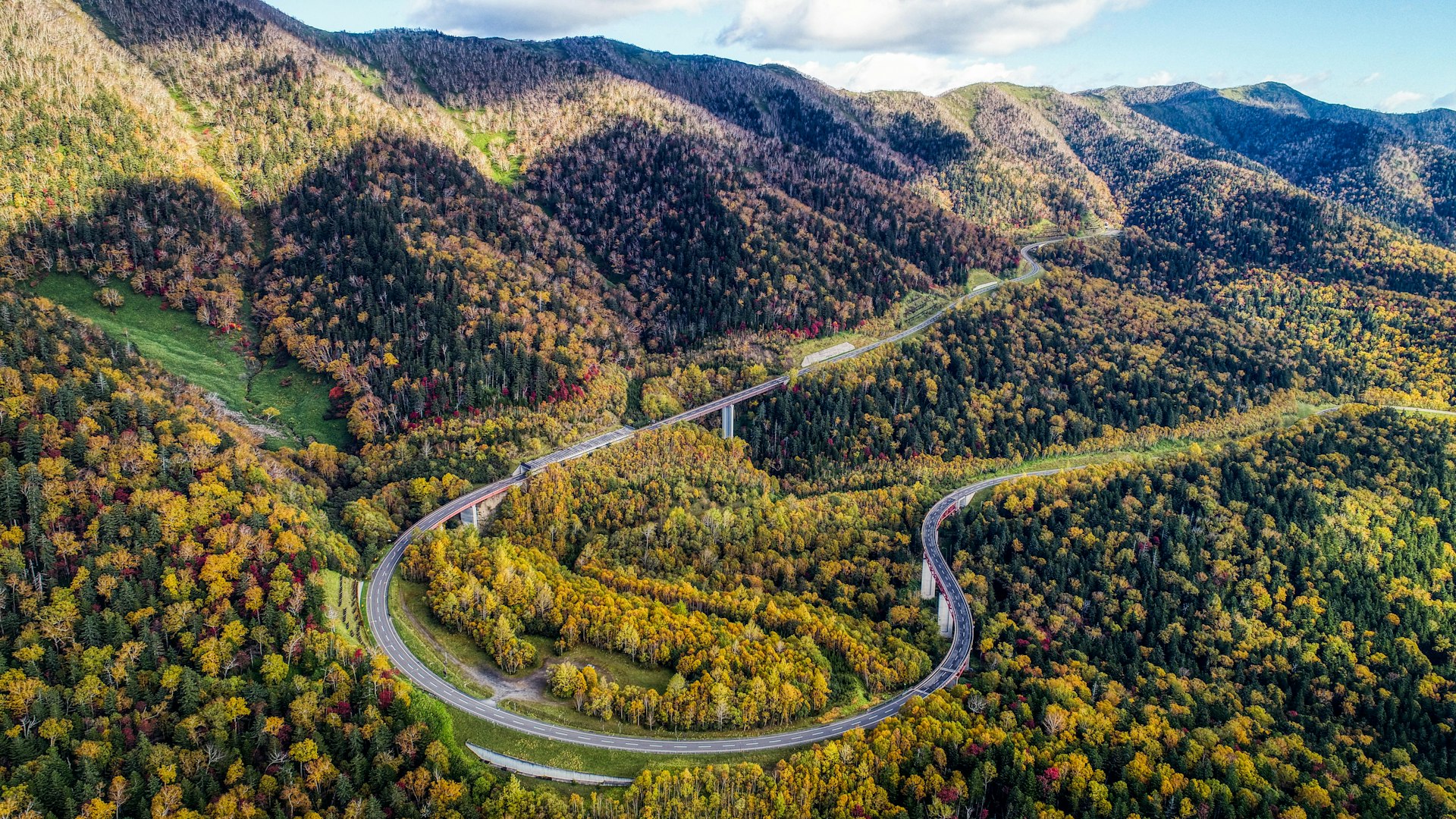 An aerial shot of Mikuni pass in Hokkaido. The image shows a road, which is traffic free, curving through a green forest.