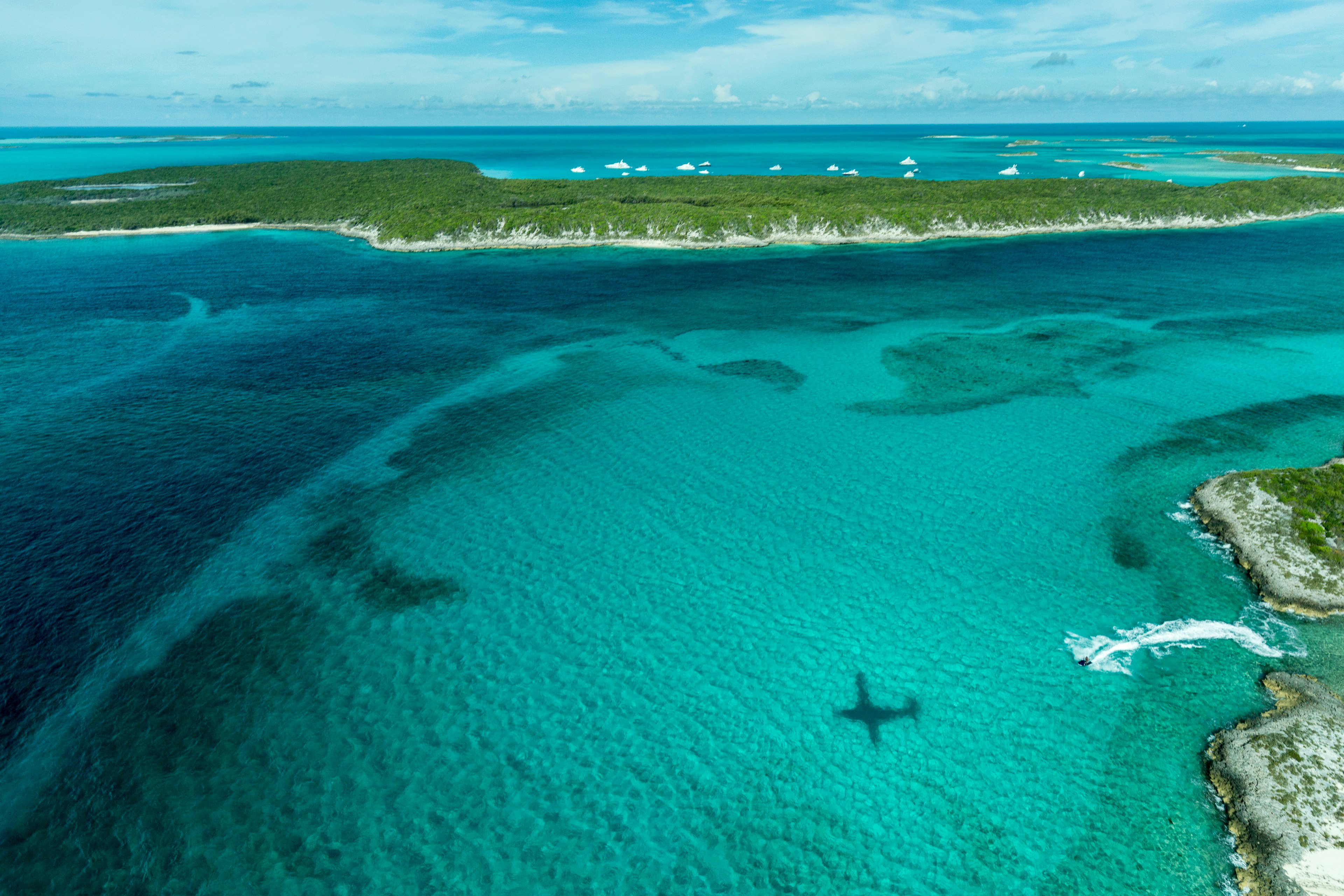 Aerial looking down at the airplane's shadow, a jet ski and clear tropical water and islands in the Exuma Chain of islands the Bahamas near Staniel Cay.