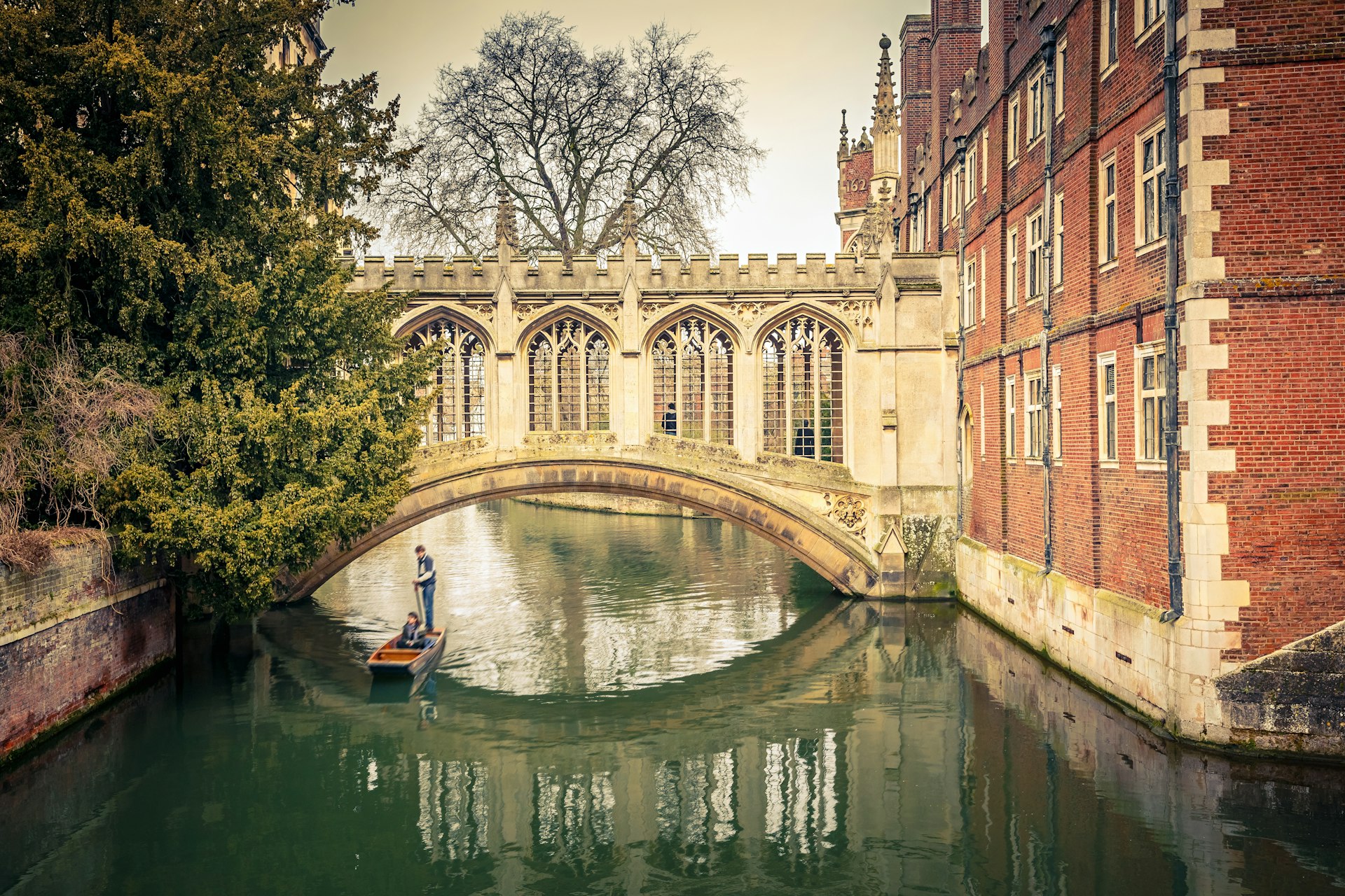 A quaint stone bridge over a waterway linking two buildings. A narrow boat with a pilot standing at the back of it passes nearby 