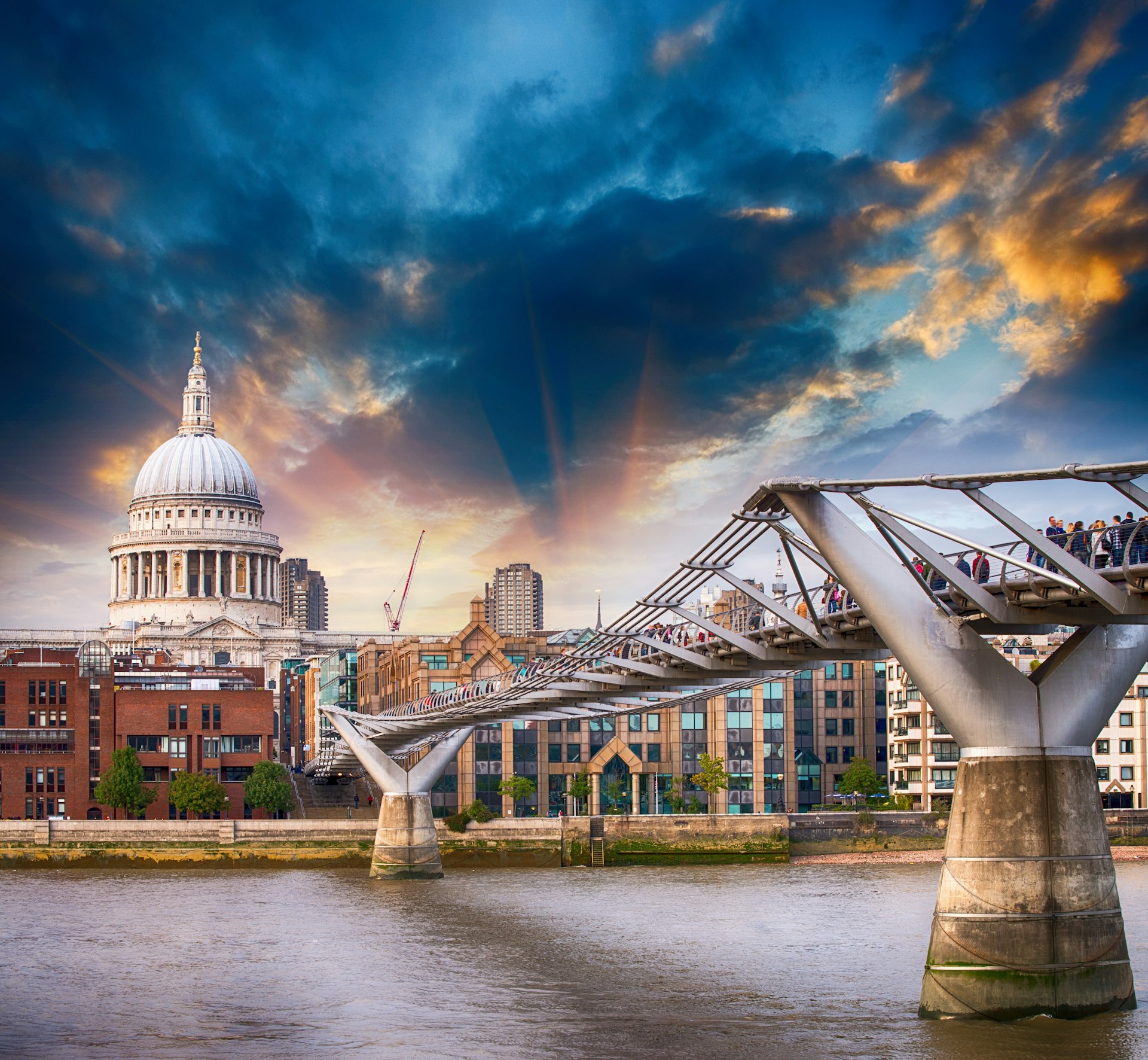 A metal footbridge spans a wide river, leading to a domed church building. There's an atmospheric sky of dark clouds and sunlight.