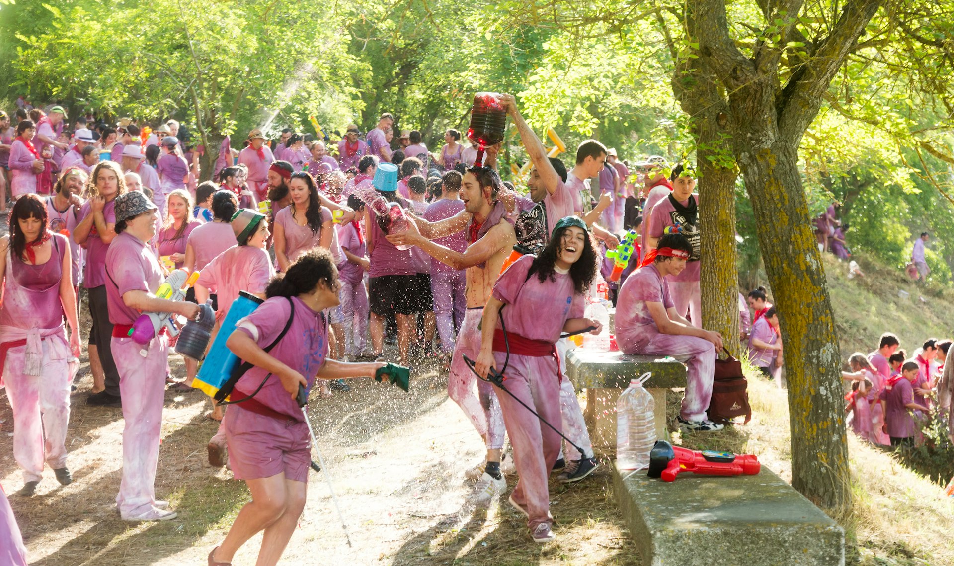A crowd of people wear white clothes that are stained red by red wine. Many are laughing as wine is poured over them