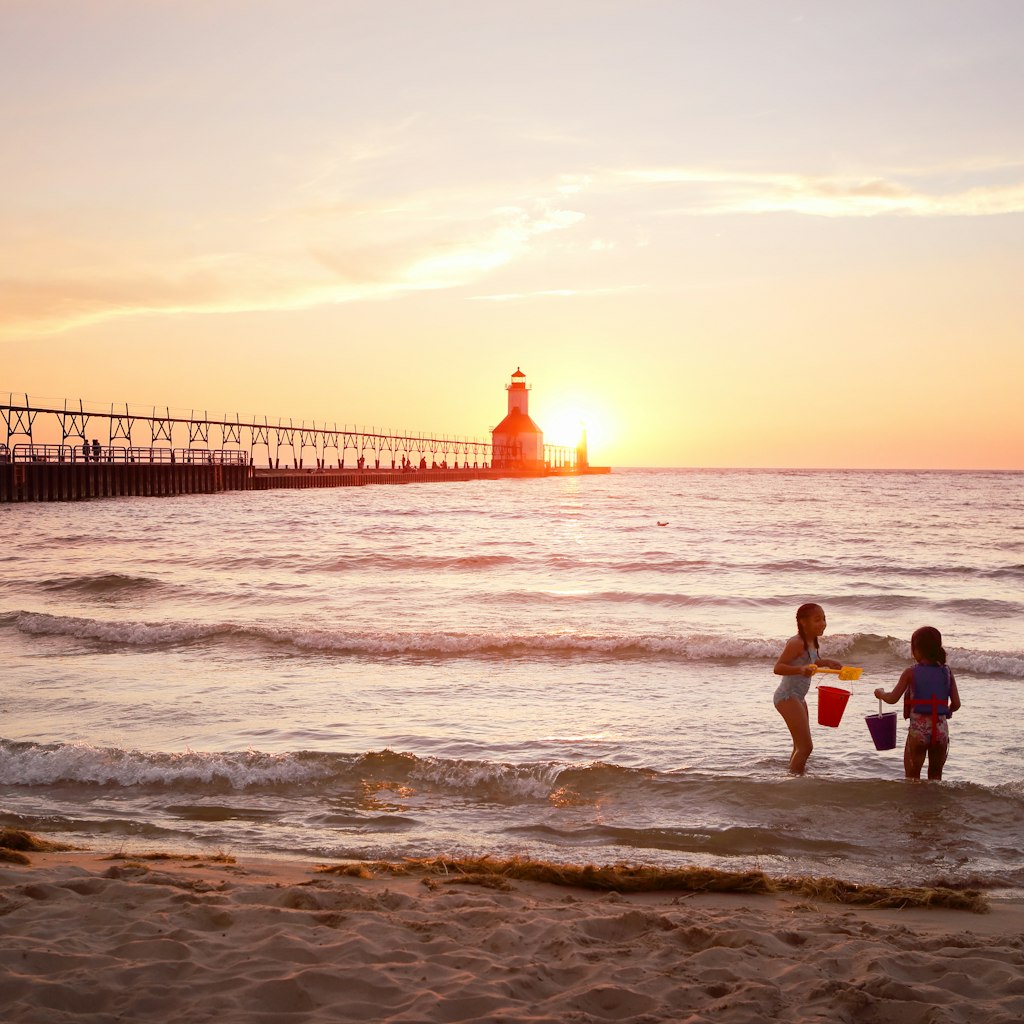 Kids playing in the water in front of St Joseph Lighthouse on Lake Michigan during sunset.