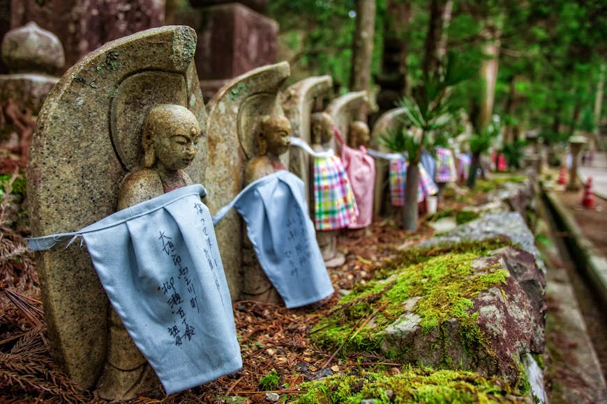 Okunoin-kyrkogården i Koyasan, Japan.  Ett antal små stenstatyer är inslagna i tyg, mitt i en skogsmiljö.