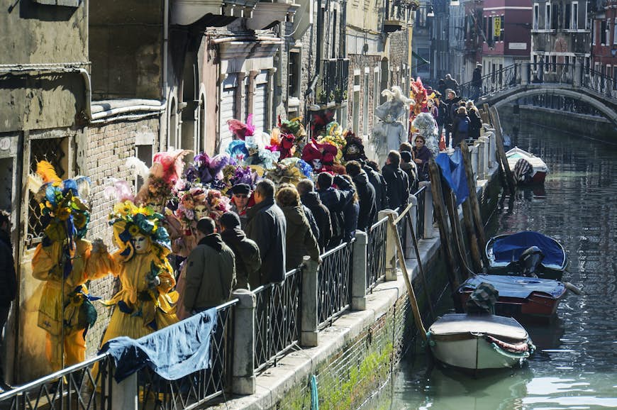 En karnevalståg med människor klädda i ljusa kostymer längs en smal kanalväg i Venedig. 