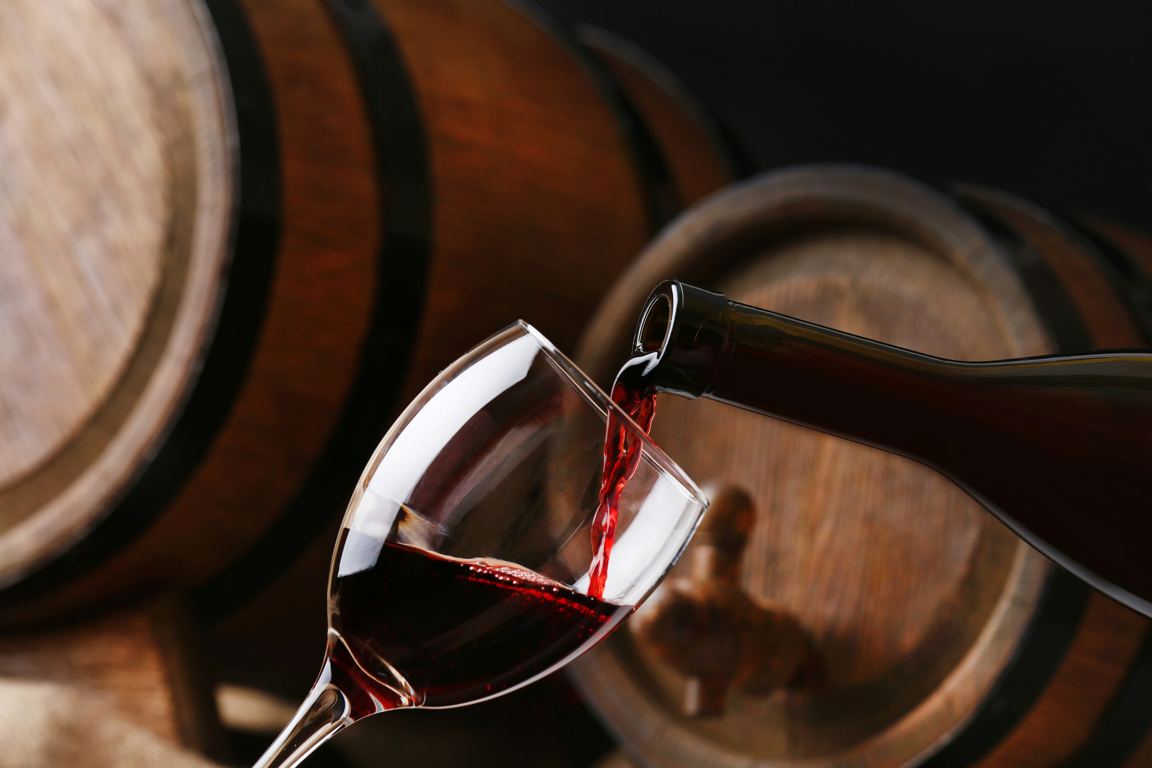 Pouring red wine from bottle into glass with wooden wine casks in the background