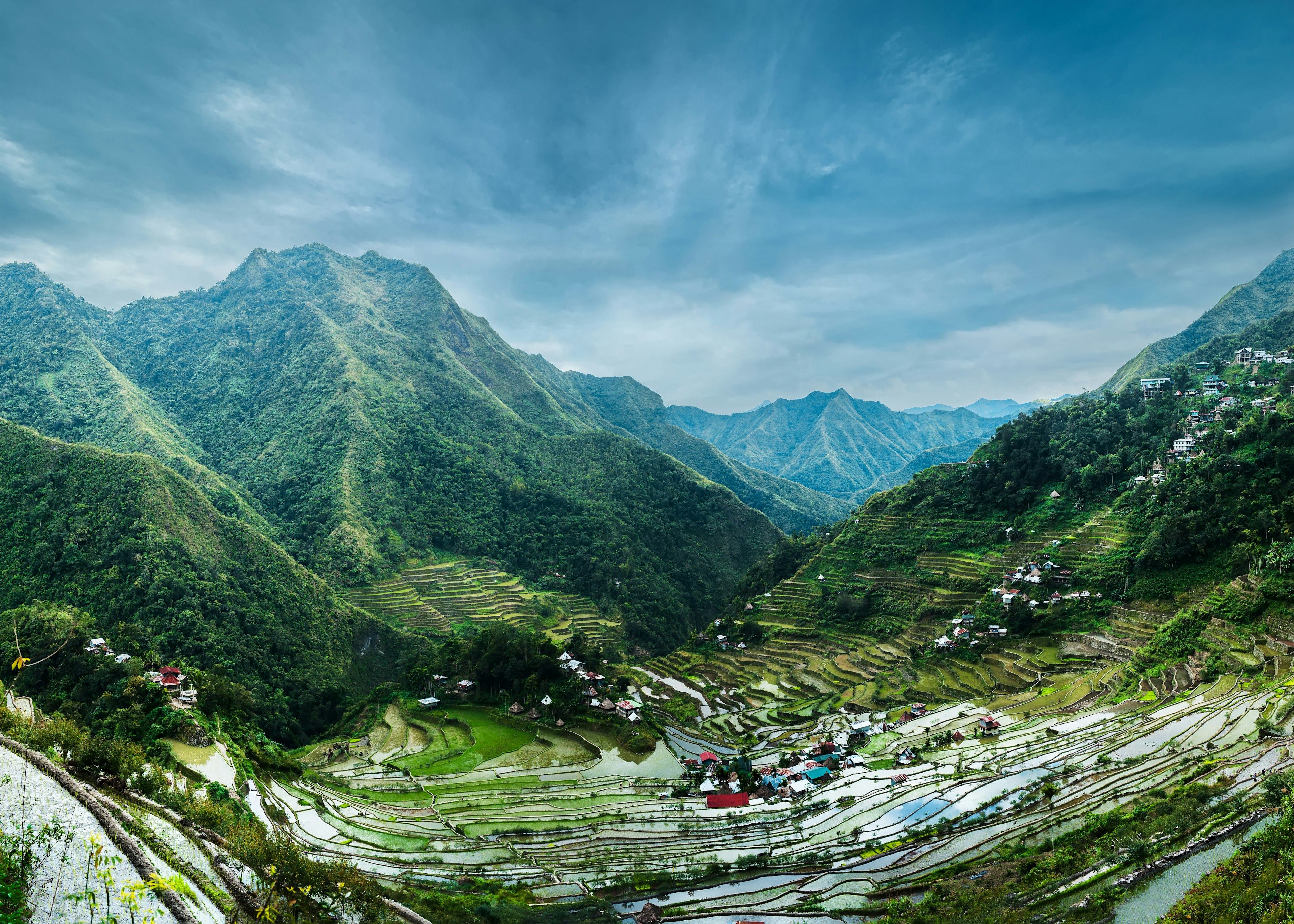 A series of rolling green rice terraces in Ifugao province, Luzon, the Philippines. The fields are surrounded by mountains beneath a cloudy blue sky.