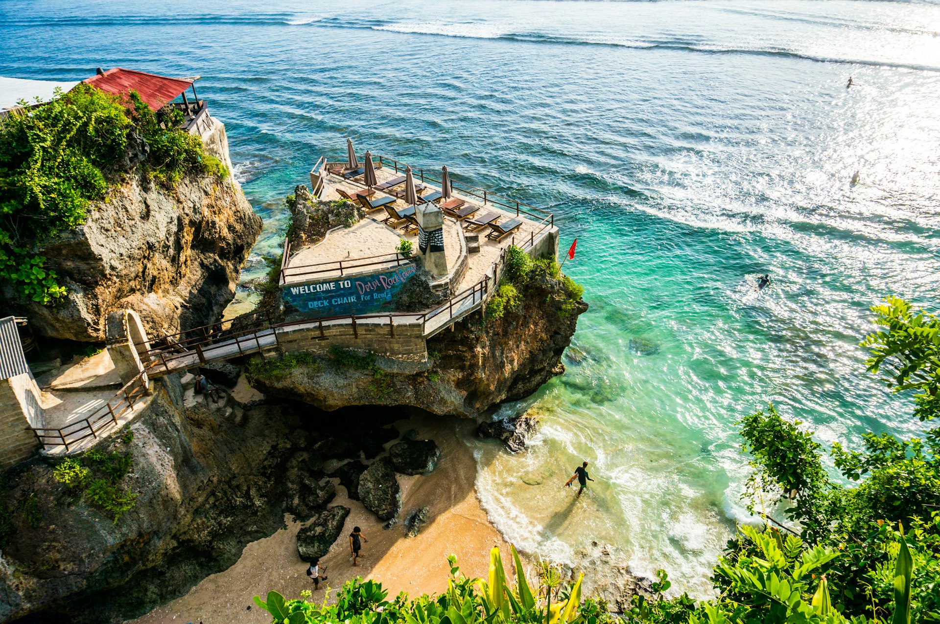 A surfer walking out to the ocean on Suluban Beach, near a sheer cliff with a lounge on top