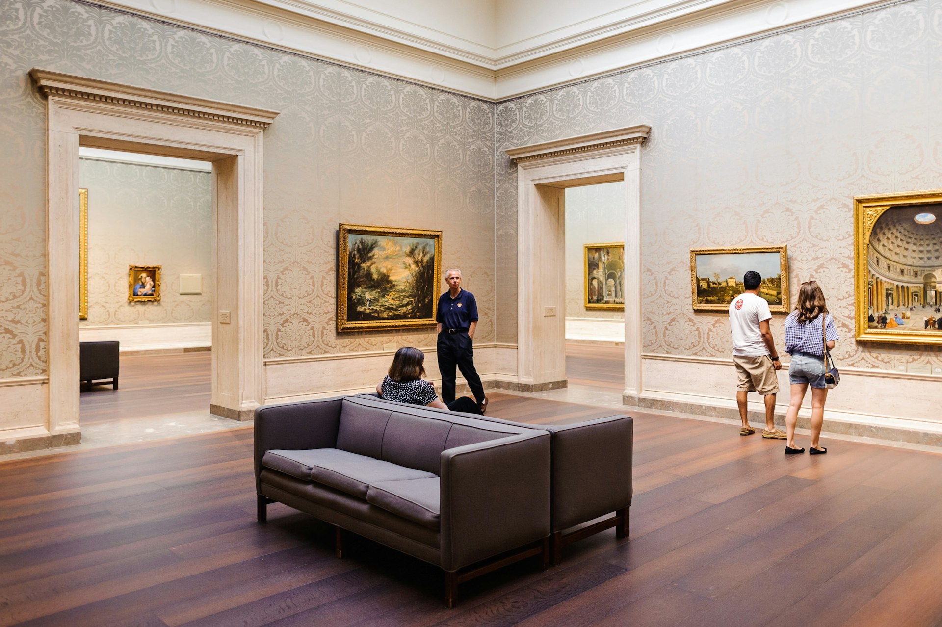 The interior of an art gallery. There is couch in the middle of the room and a few people viewing paintings on the wall.