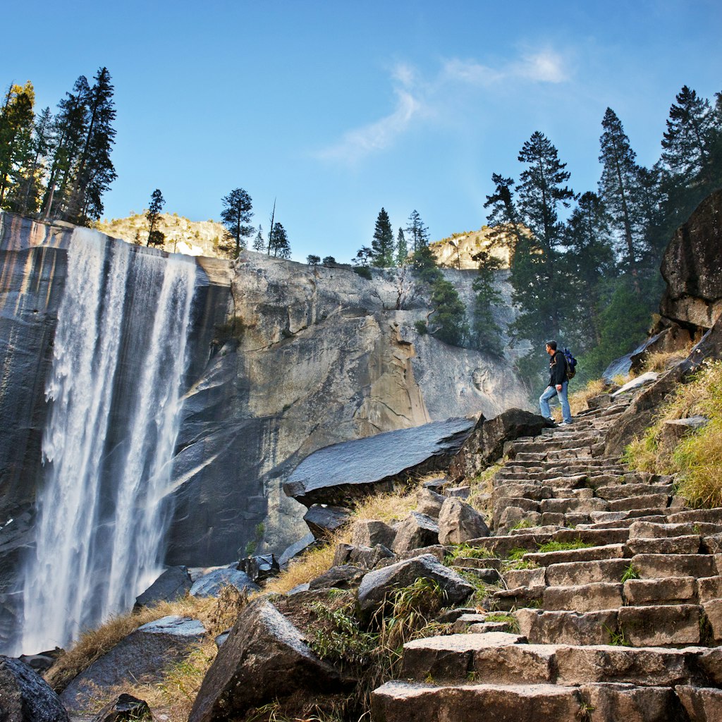 A male hiker standing on stone steps and admiring a waterfall in Yosemite National Park.