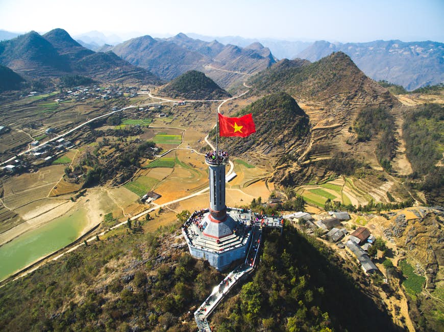 An aerial view of Lung Cu flag tower in Ha Giang, Vietnam. The large flag stands on top of a mountain, with views over the surrounding green valleys and hills.