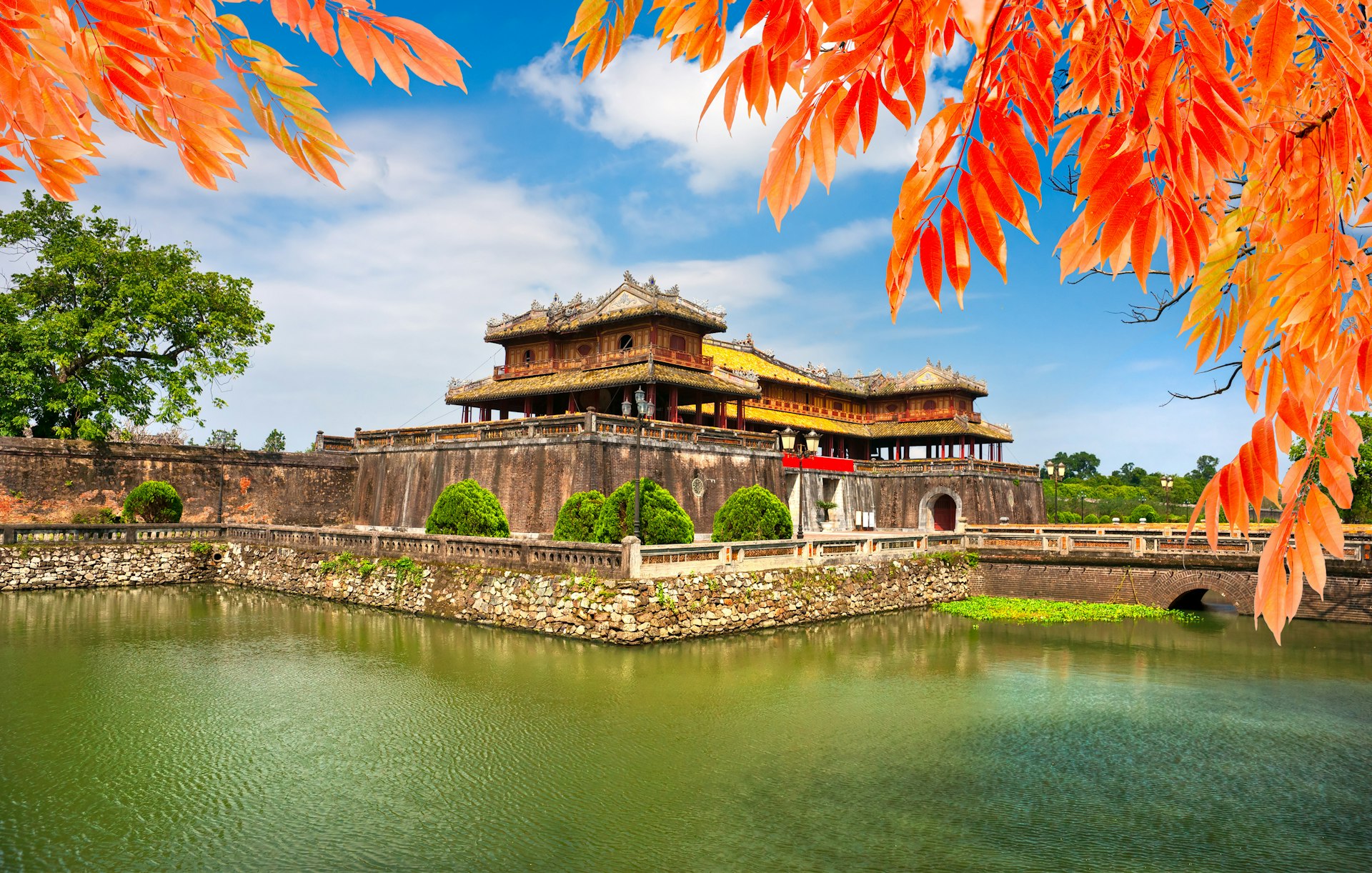 A view of the entrance of the Citadel in Hue, an ancient walled enclosure within the city housing temples and palaces. The stone structure has a moat around it, and the image is framed by red leaves of a nearby tree.