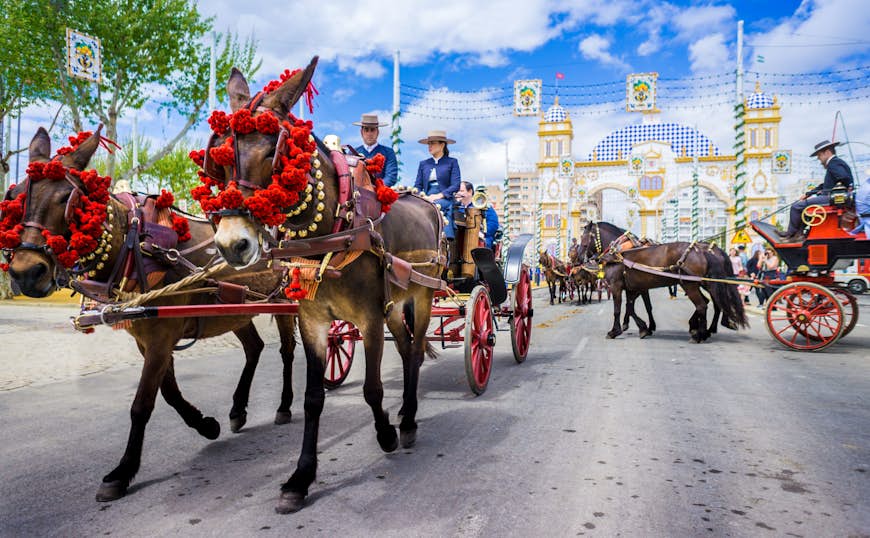 Horse-drawn carriages pass through a square. People are in traditional dress celebrating a festival