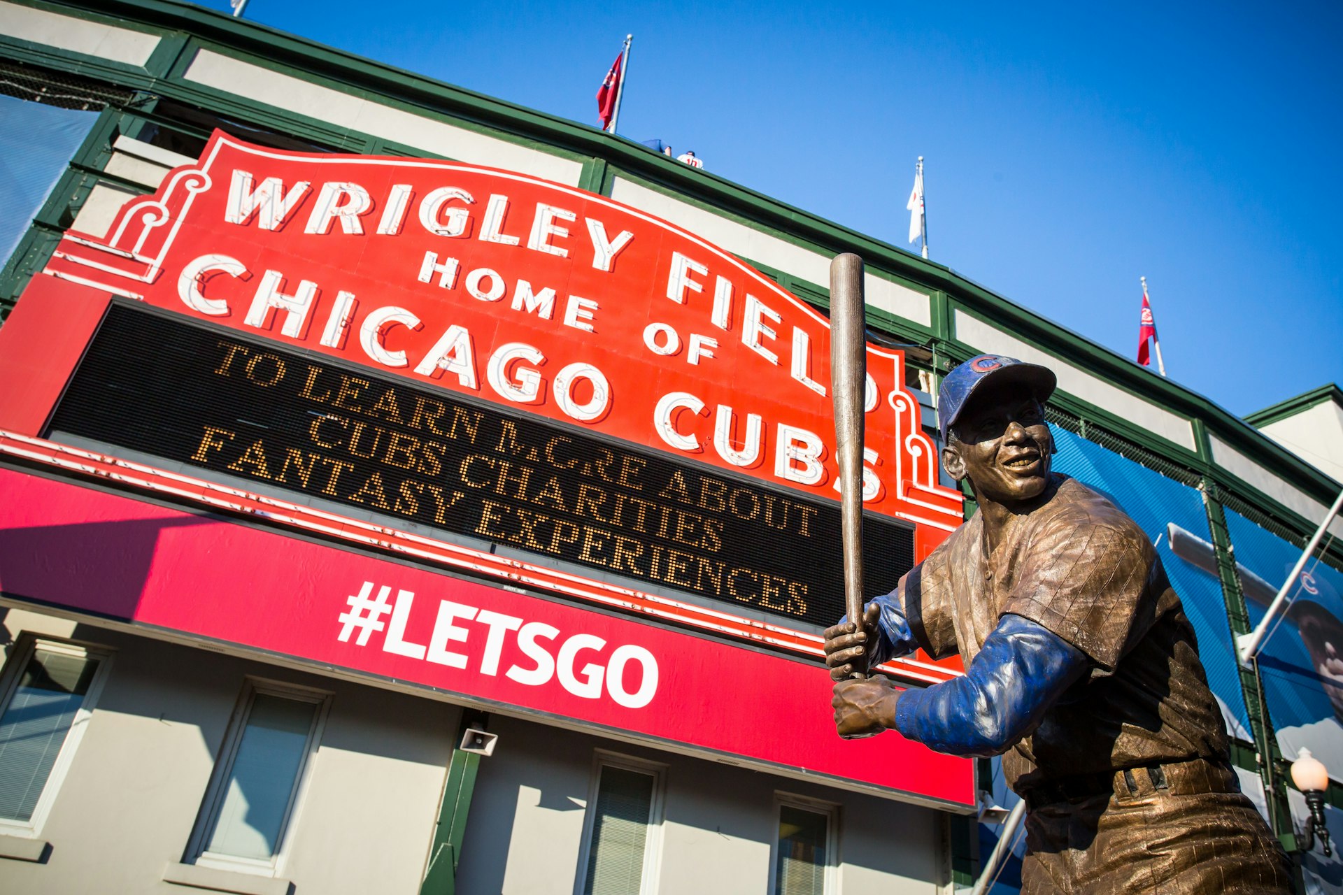 A large red-and-white sports sign outside a stadium with a bronze statue of a baseball player in front of it