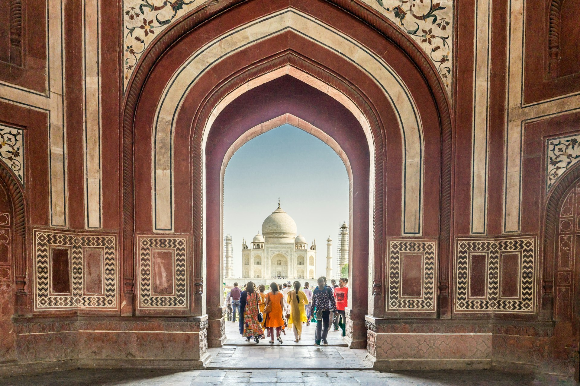 People in colorful clothes pass through an arched doorway. Beyond them is a great white marble dome of the the iconic Taj Mahal