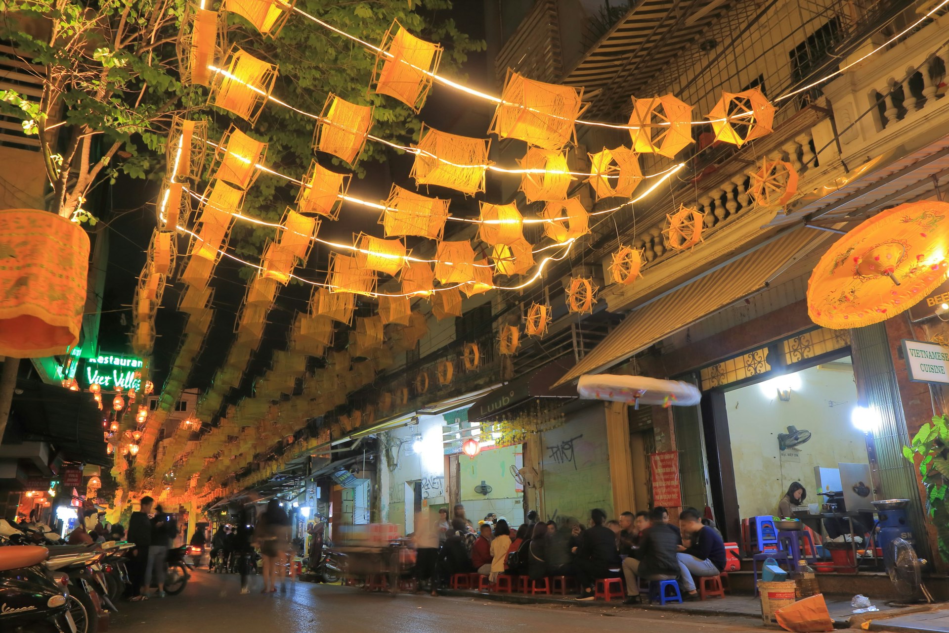 People sit outside bars and restaurants in Hanoi's Old Quarter. It is night and tables and chairs have been set out on the road outside the businesses.