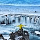 A man sits in front of Godafoss Waterfall surrounded by snow.