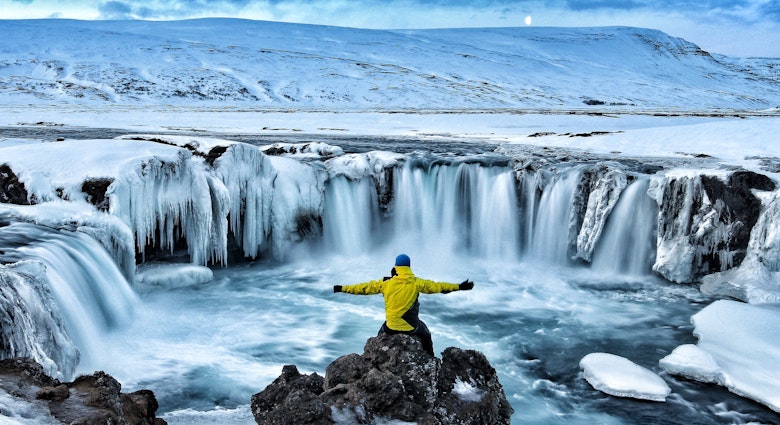 A man sits in front of Godafoss Waterfall surrounded by snow.