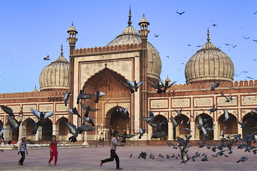 Το Τζαμά Μαστζίντ στο Δελχί, ένα από τα μεγαλύτερα τζαμιά στην Ινδία. Ένα μικρό παιδί κυνηγάει περιστέρια στην αυλή του τζαμιού, που πετούν στον ουρανό. Πίσω του βρίσκονται οι τρεις εμβληματικοί τρούλοι του τζαμιού στην οροφή του μεγάλου κτιρίου.