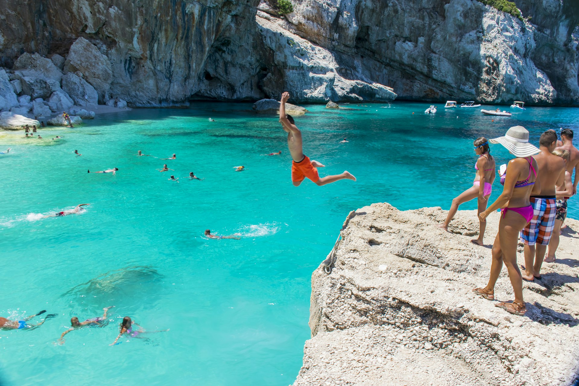 People jump off rocks into the turquoise waters of the sea below