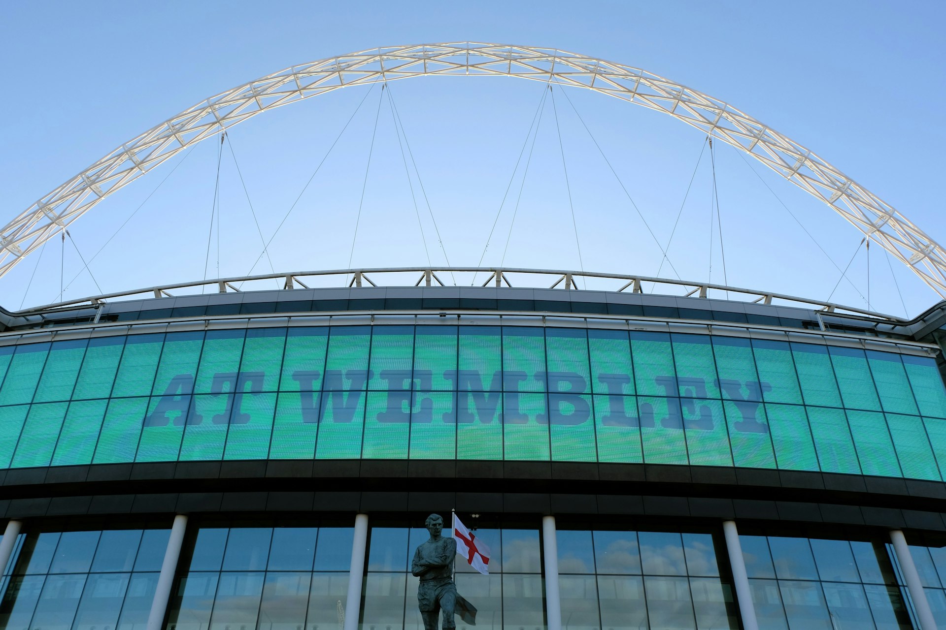 A large series of glass windows on the upper part of a large stadium. A huge metal arch rises over the top. 
