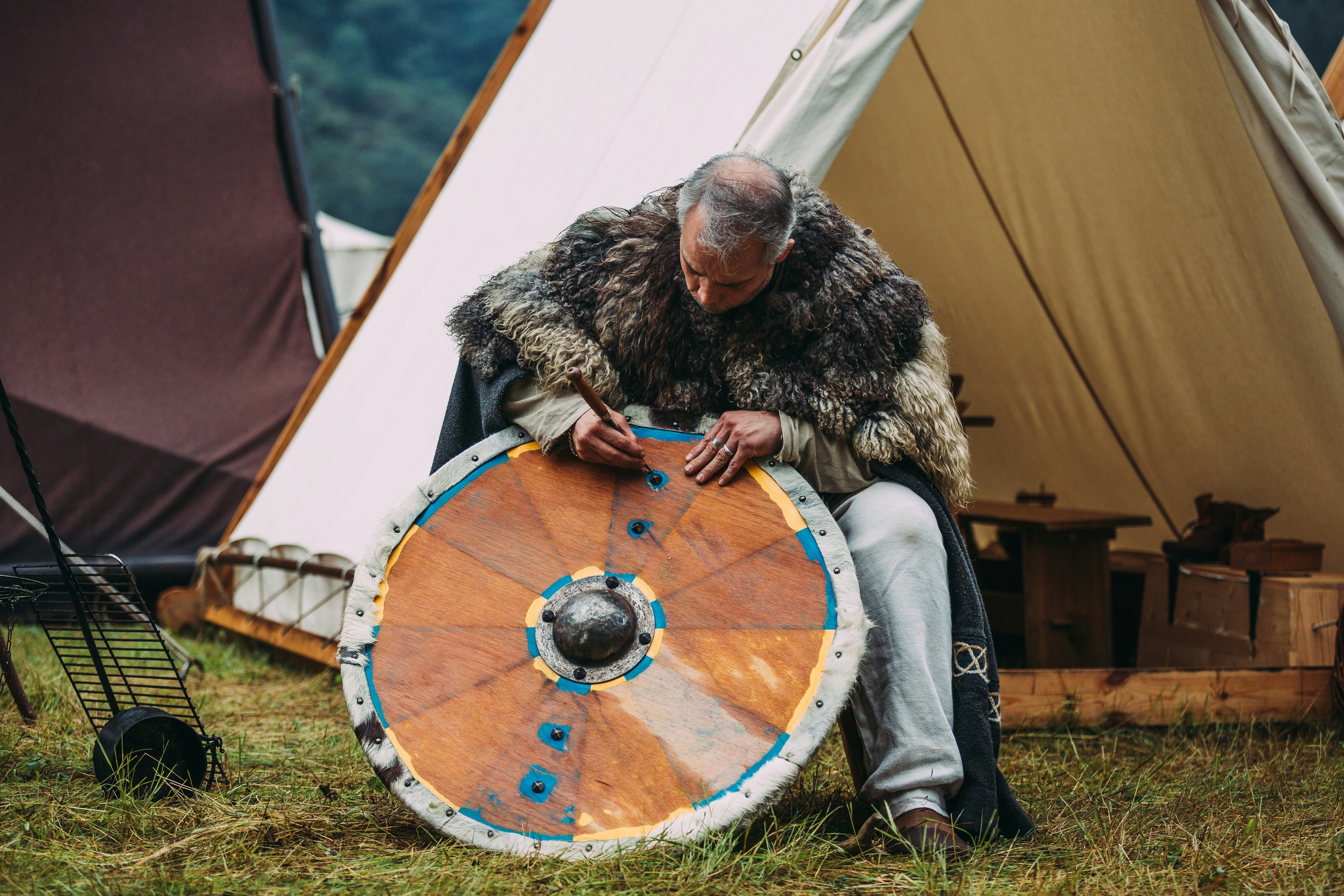 A man dressed in fur sits and leans over a traditional Viking shield