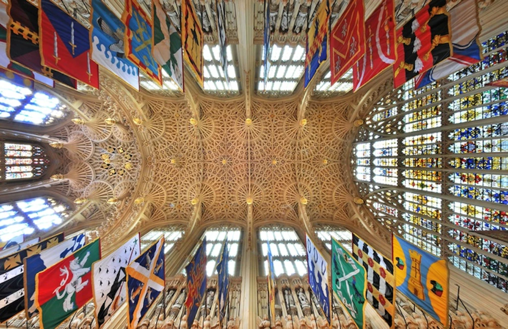 A shot straight up towards a ceiling with intricate stone webbing across it. There are colorful flags hanging around the walls