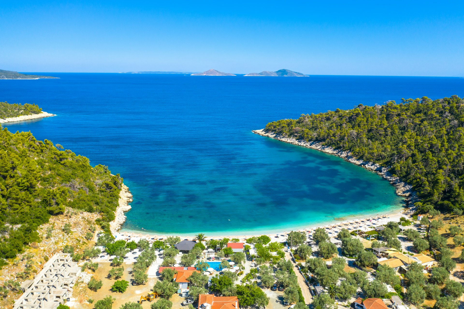 An aerial view of a white strip of sand and blue waters off the coast of Alonnisos Island, Greece. The beach is backed by a small village, with numerous buildings.