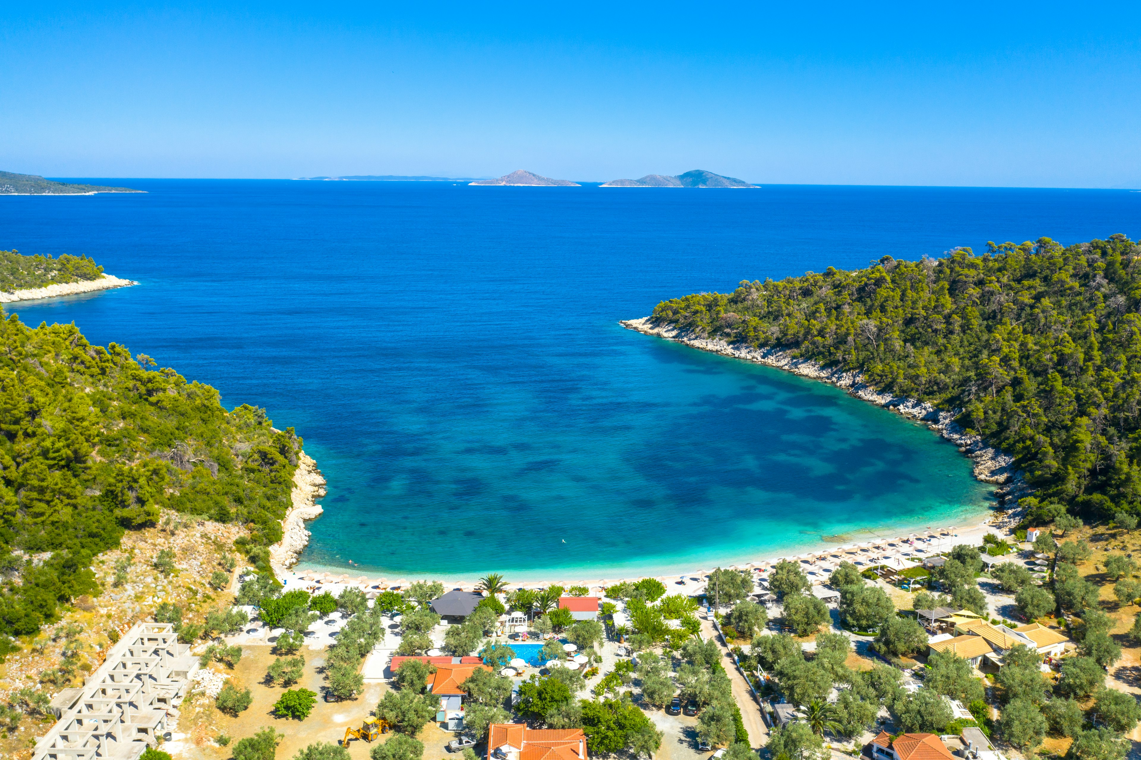 An aerial view of a white strip of sand and blue waters off the coast of Alonnisos Island, Greece. The beach is backed by a small village, with numerous buildings.