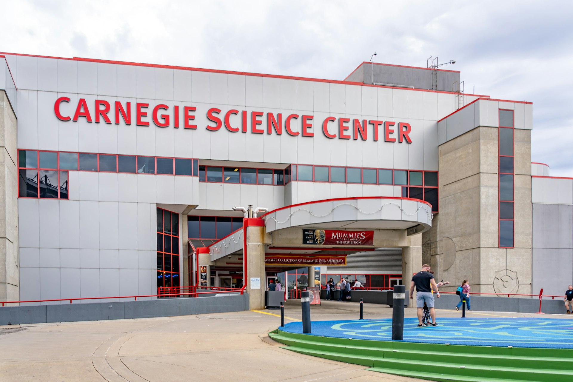 Entrance of Carnegie Science Center in Pittsburgh, Pennsylvania There are a few people standing outside of the red and gray building