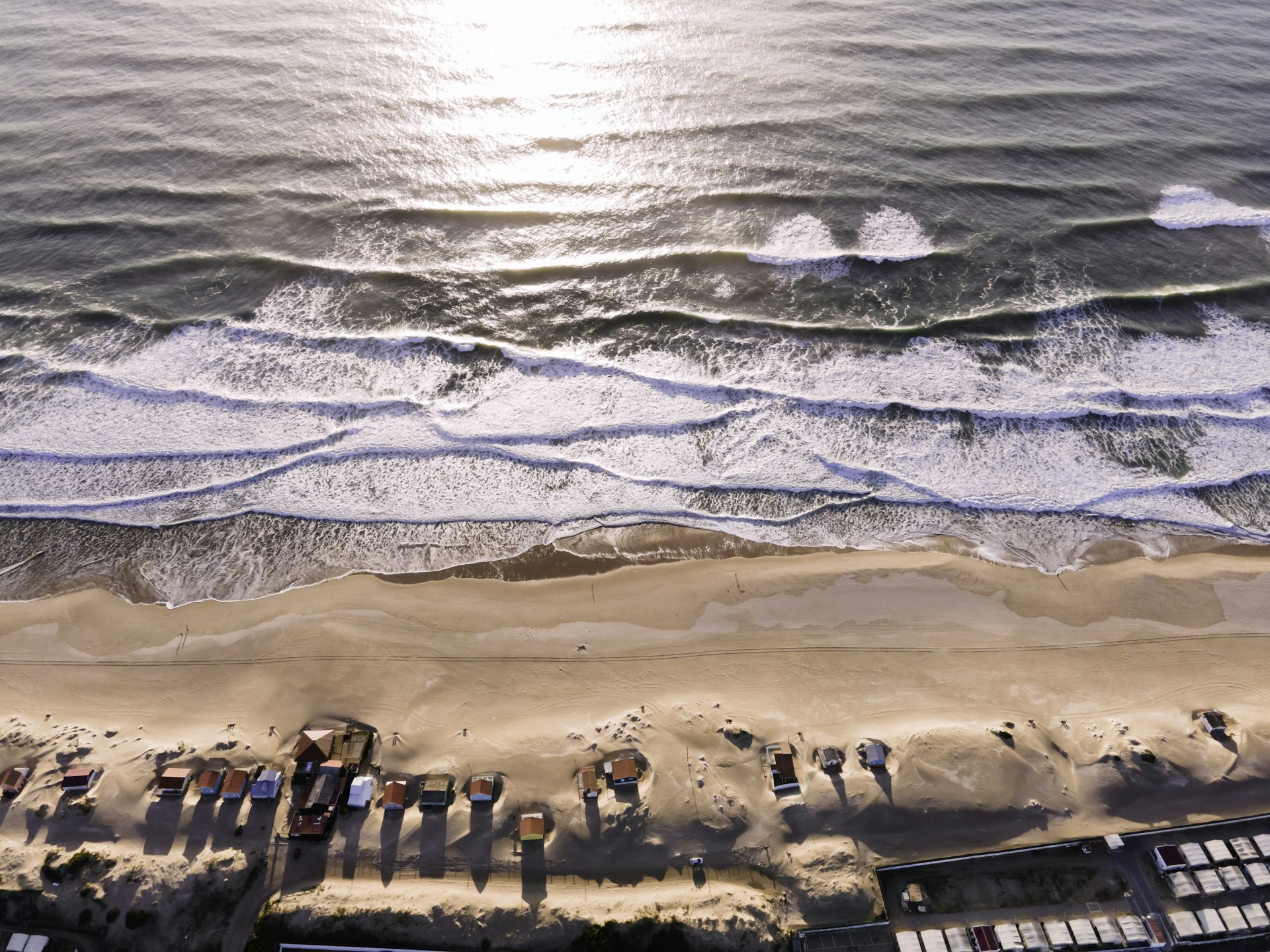 Aerial view of waves breaking on the long, beautiful beach of Costa da Caparica. A number of small shacks stand on the otherwise empty sand.