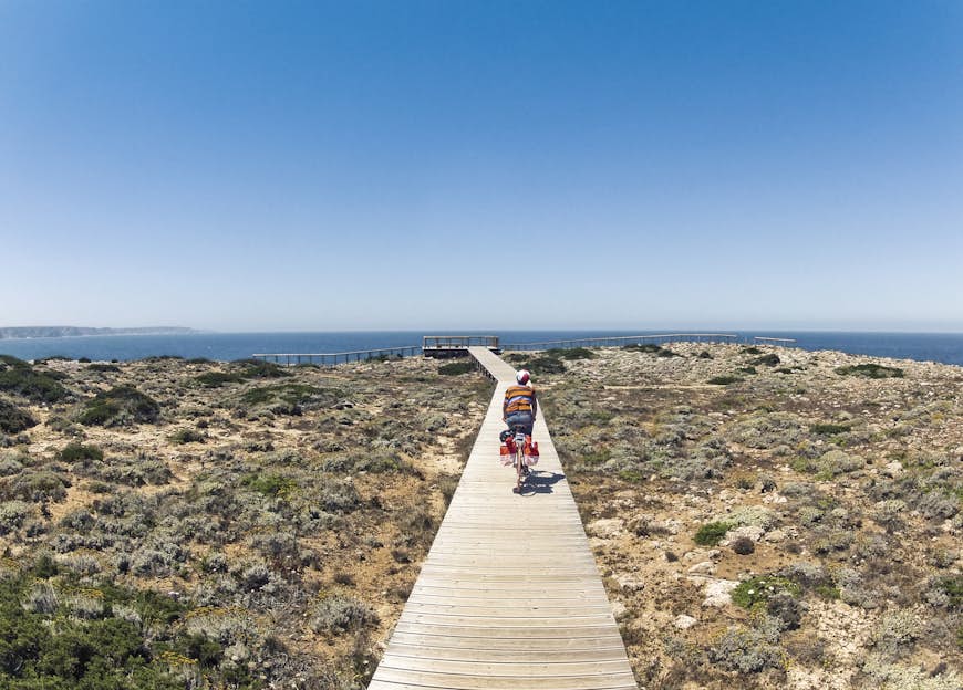 A young woman riding a bicycle on a narrow wooden boardwalk in Carrapateira, Portugal. In the distance the blue sea is visible.