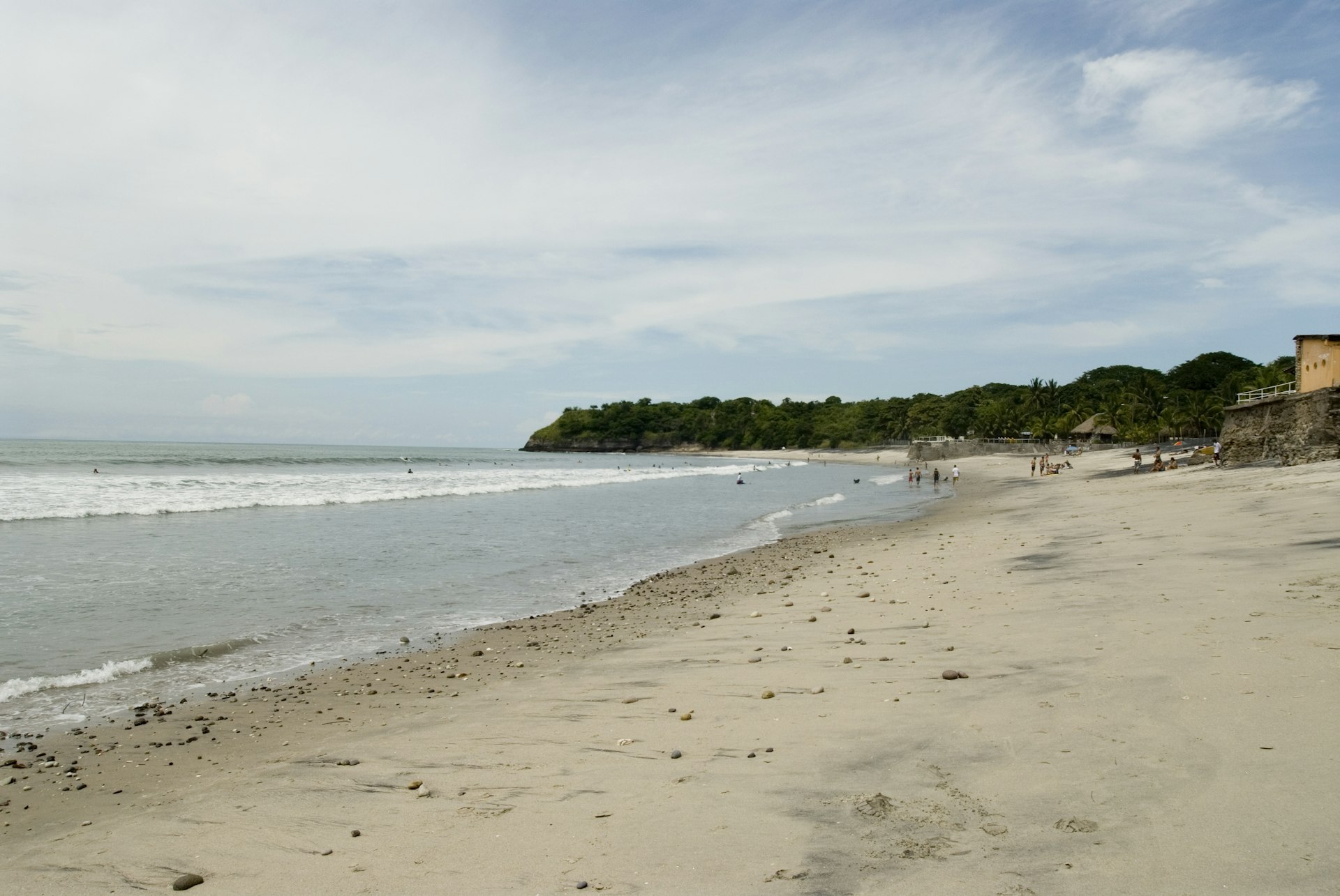 A wide angled view of El Palmar Beach in Panama. The beach is sandy and gently slopes down into a gentle sea. A few families stand around on the sand in the distance.