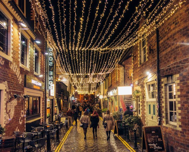 A pedestrianised cobbled street at night time, with fairy lights strung high up and people enjoying a night out  