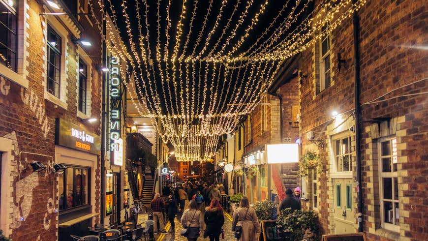 A pedestrianised cobbled street at night time, with fairy lights strung high up and people enjoying a night out  