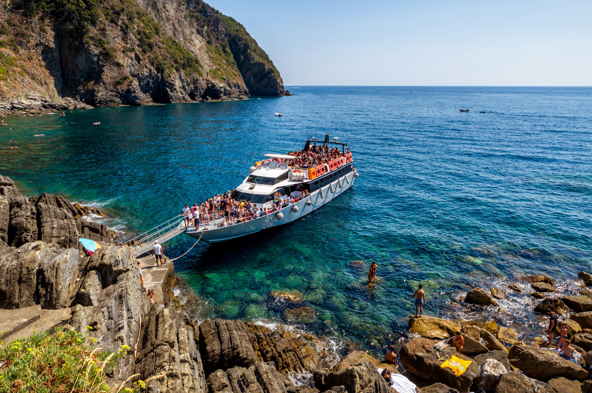 A ferry boat floats in the shallow blue waters off the coast of Riomaggiore in Cinque Terre. The boat is very busy with many people up on deck.