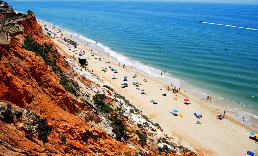 En översikt över Praia da Falesia i Portugal.  Den tunna remsan av gyllene sand är full av människor som solar.  Bakom sanden finns ljusbruna klippor och framför ett turkost hav.