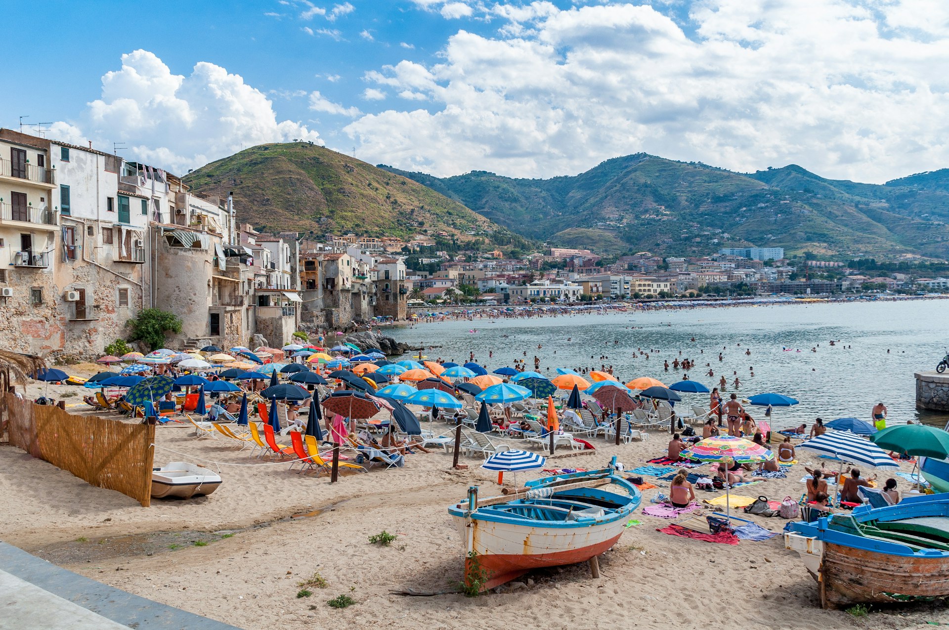 Fishing boats are marooned on the sands of Cefalu Beach in Italy. Behind them are a number of beach umbrellas where beach goers lie on the sand. In the background, the sea is visible, as are some buildings from a historic-looking port town.