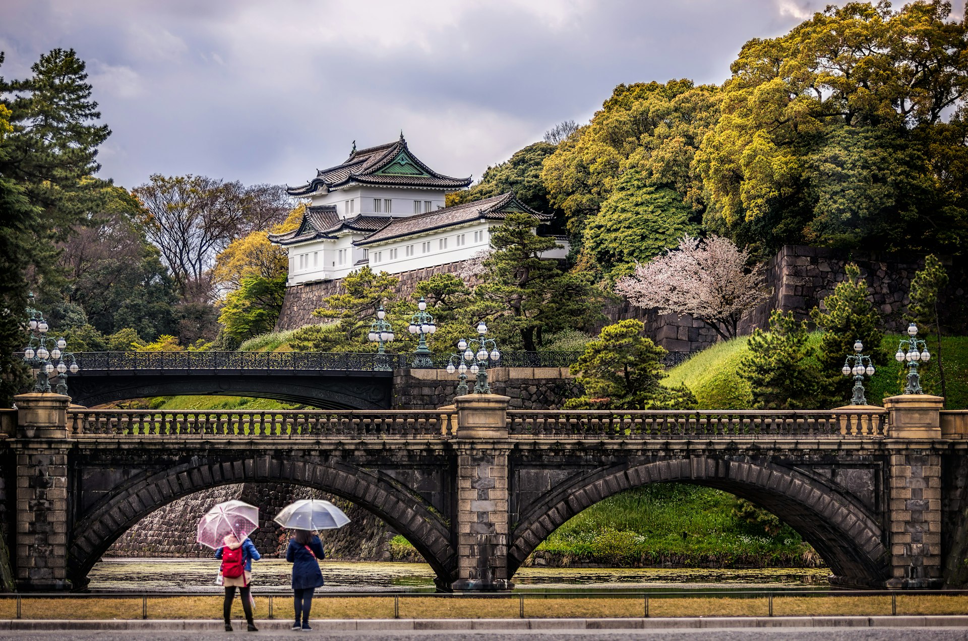 Two women stood beneath umbrellas stand in front of a bridge at the Imperial Palace in Tokyo