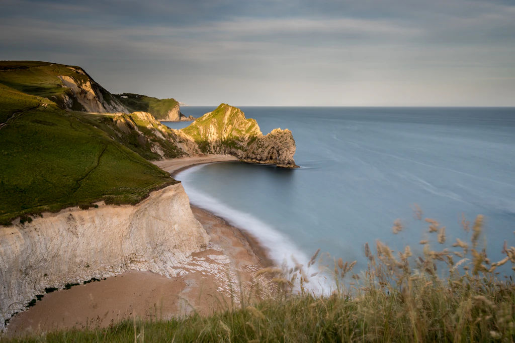 Durdle Door Undulating cliffs leading down to a sandy and shingle beach, with a huge stone archway at one end