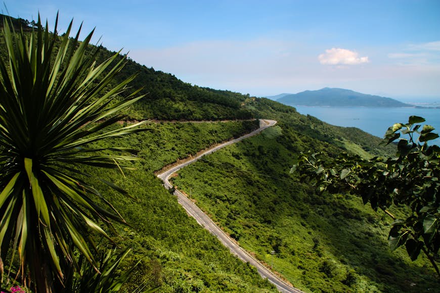 A road curls around a green mountain as part of the Hai Van Pass in Vietnam. In the background a blue ocean is visible.