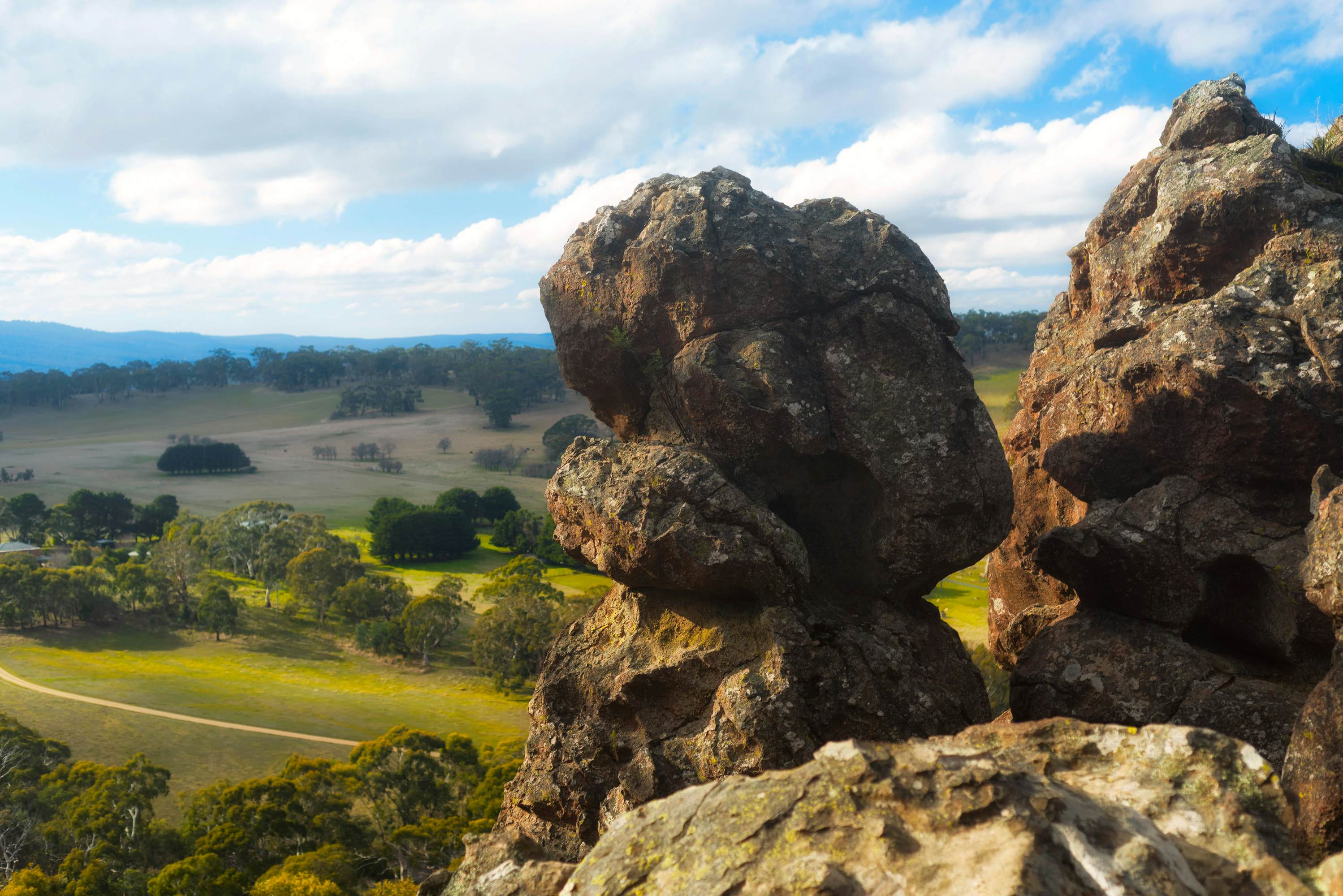 A oddly-shaped boulder stands on a hilltop at Hanging Rock, Australia. The area is a sacred site for the Wurundjeri people. In the distance, far below, forests and fields are visible.