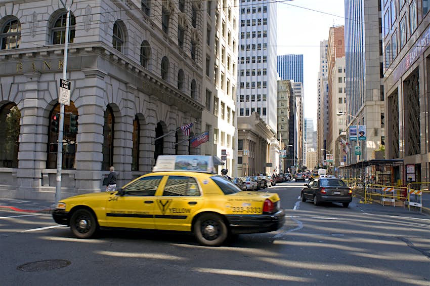A yellow taxi drives through the skyscrapers of the Financial District in San Francisco.