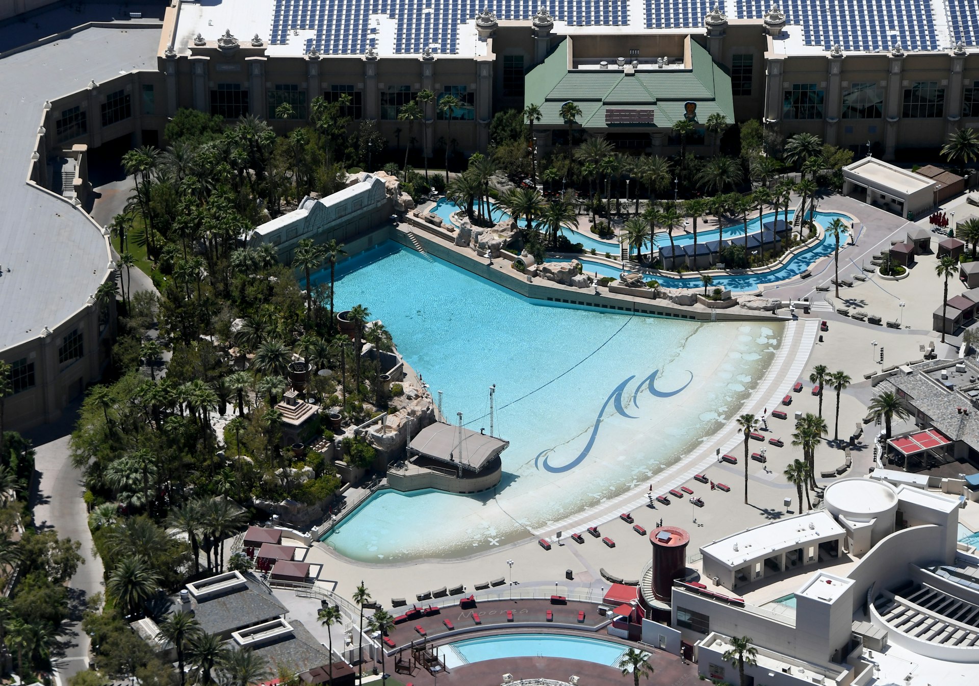 Aerial view of Mandalay Bay's manmade beach, which is a large fan-shaped pool. It's surrounded by palm trees and poolside cabanas. The back portion of the Mandalay Bay can also be seen. 