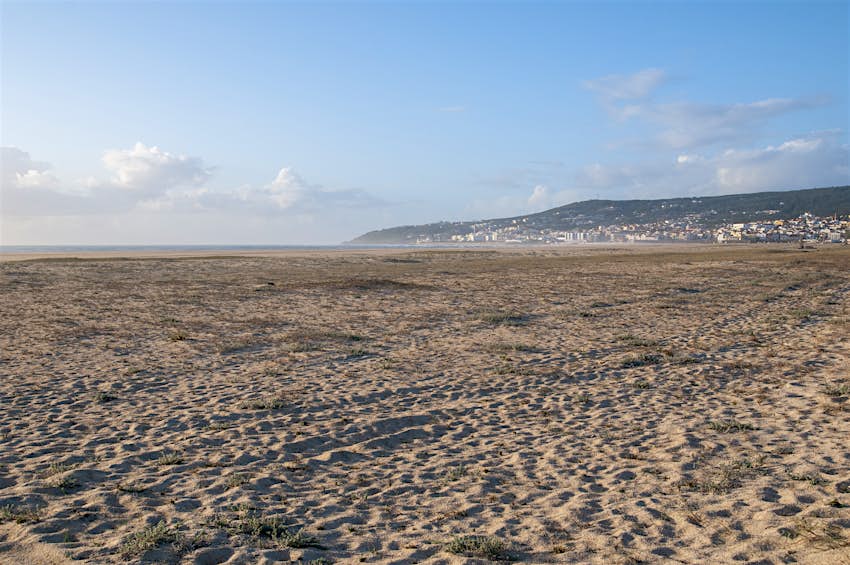 Praia da Ficura da Foss Foto de baixo ângulo de uma grande praia, visível ao mar ao longe, ao lado, dois edifícios. A majestosa praia é dourada e vazia.