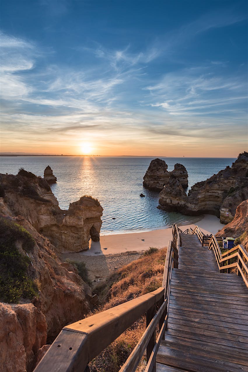 Praia do Camilo (Priya do Camilo) ao nascer do sol em Lagos, Algarve, Portugal Vista vertical dos degraus de madeira que conduzem à Praia do Camilo, Portugal ao nascer do sol. A pequena enseada de areia é cercada por rochas atmosféricas e está vazia de pessoas.