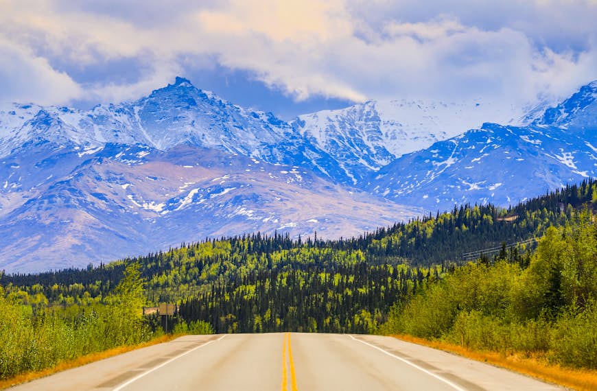 An empty road leads toward snow-capped mountains in Denali National Park, Alaska