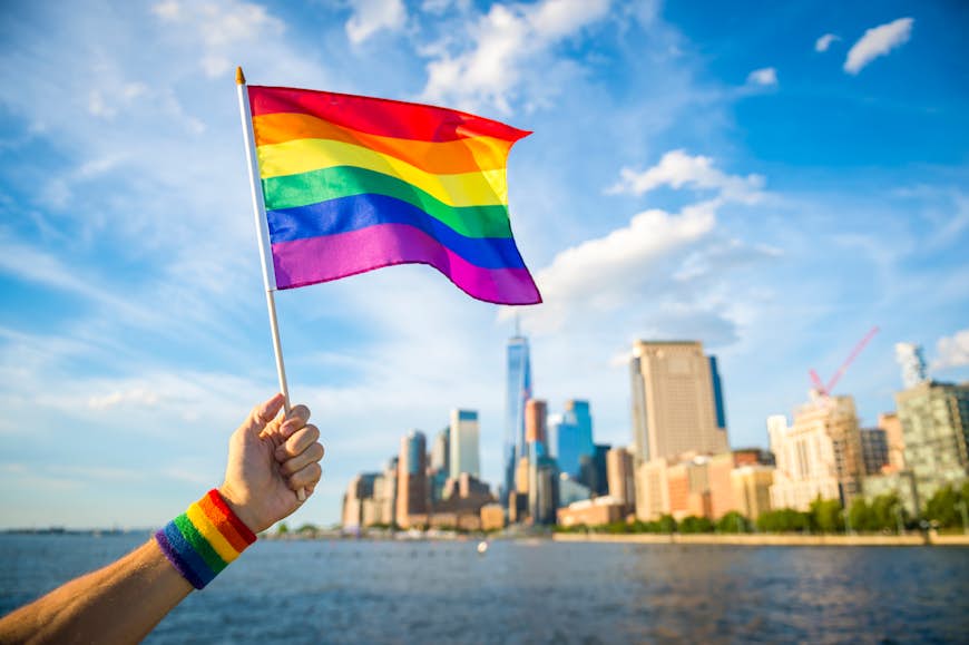 Hand holding up a colourful rainbow gay pride flag waving in the breeze with the New York City skyline in the background. 